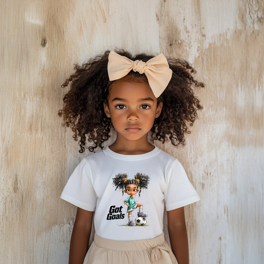 A young girl with curly brown hair wearing a large beige bow headband and a white graphic t-shirt with 'Got Goals' text, standing against a textured beige wall.