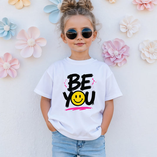 A young girl with her hair in two buns and round sunglasses wearing a kids Be You graphic t-shirt.