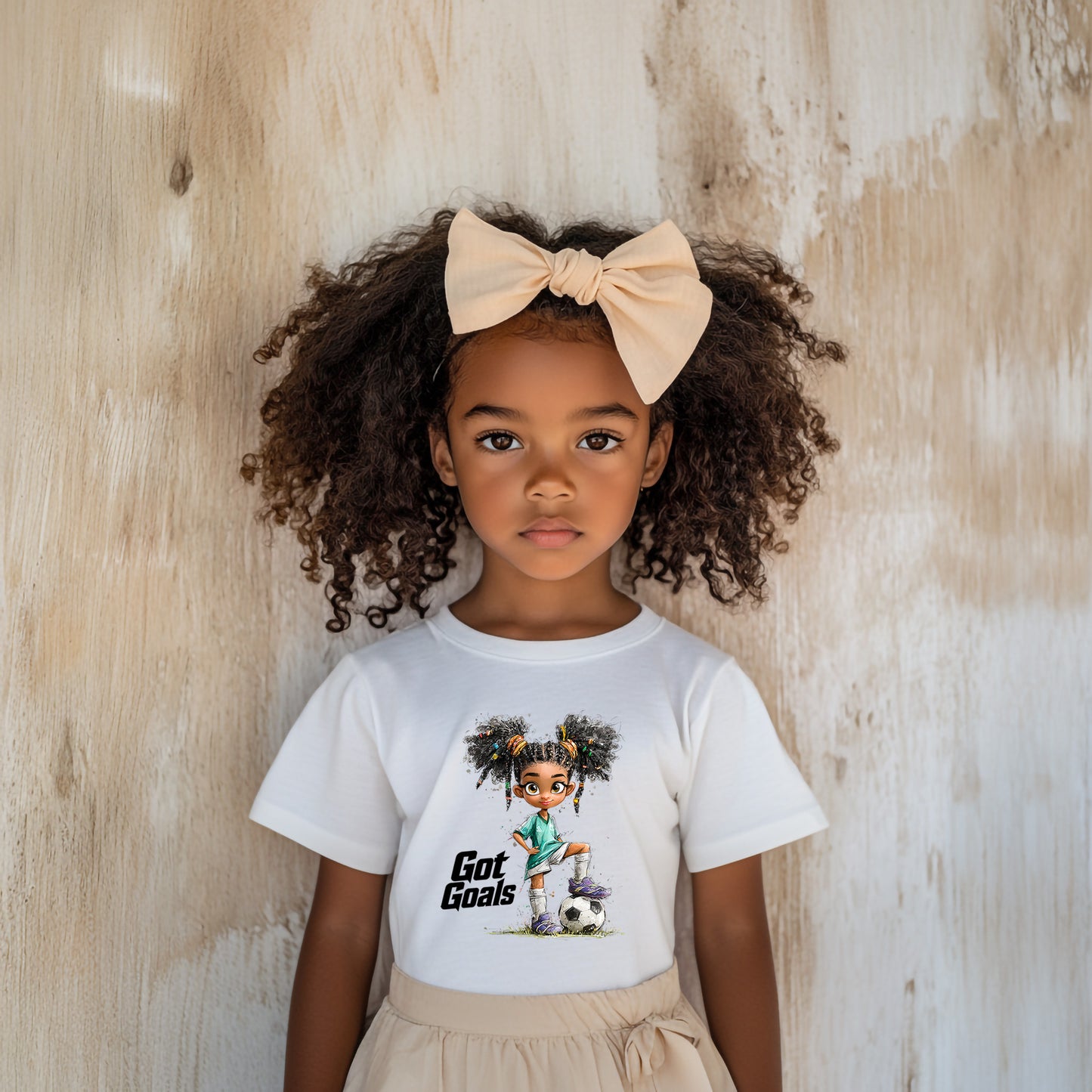 A young girl with curly brown hair wearing a large beige bow headband and a white graphic t-shirt with 'Got Goals' text, standing against a textured beige wall.
