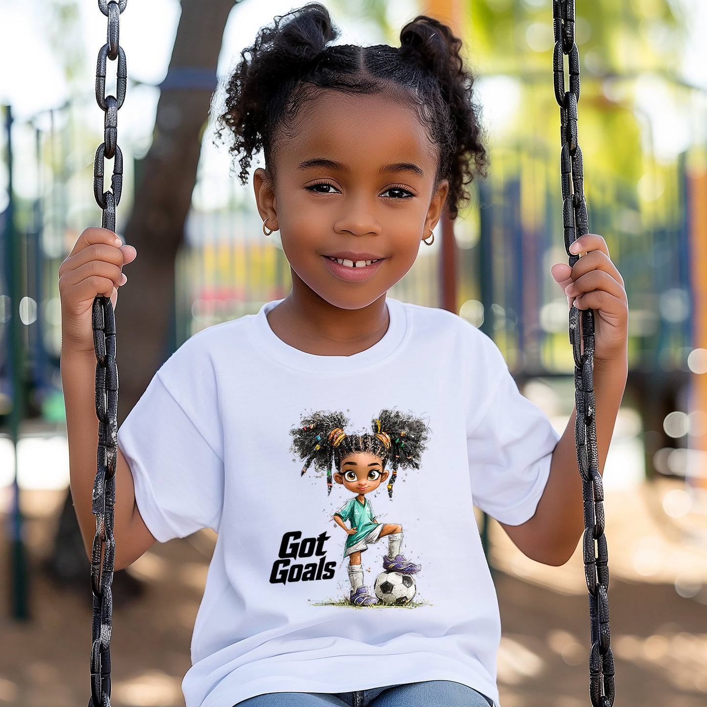 A young girl with two buns sits on a swing in a park, wearing Kids Got Goals Soccer T-Shirts