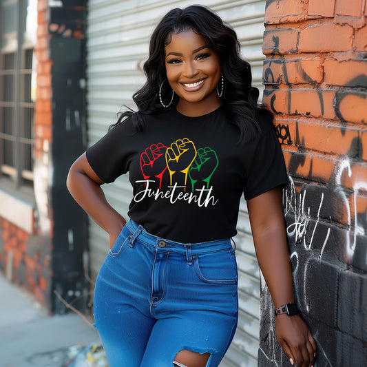Woman wearing a black t-shirt with colorful design and 'Juneteenth' text, standing against a graffiti-covered wall.