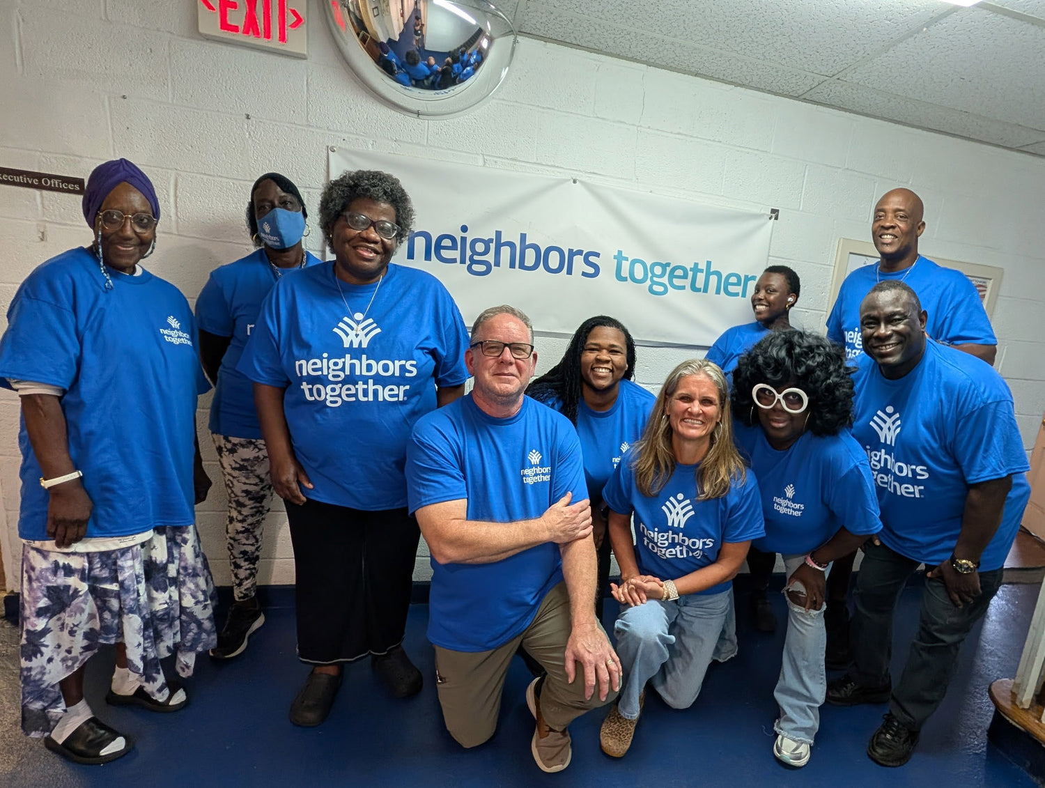 A group of employees posing together in an indoor hallway, wearing matching bright blue custom t-shirts with the white 'Neighbors Together' organization's logo printed on the front.